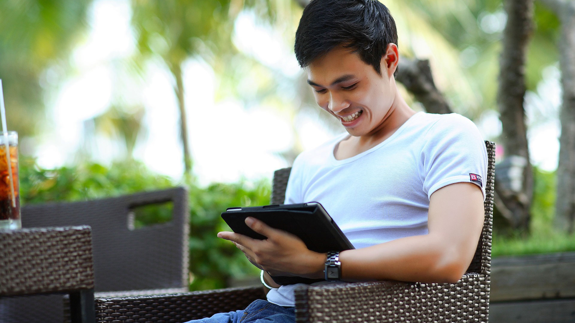 Junger Mann sitzt auf Terrasse und schaut lächelnd auf sein IPad